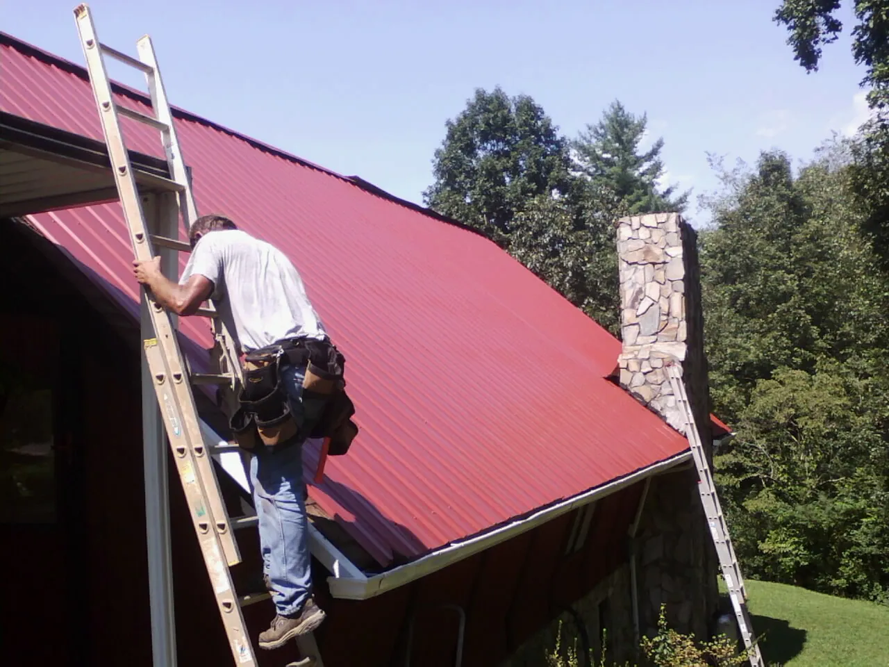 Roofer on a ladder beside a red metal roof and stone chimney in Alamo